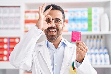 Hispanic man with beard working at pharmacy drugstore holding condom smiling happy doing ok sign...