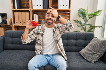 Young hispanic man with beard working on love therapy at consultation office stressed and frustrated with hand on head, surprised and angry face