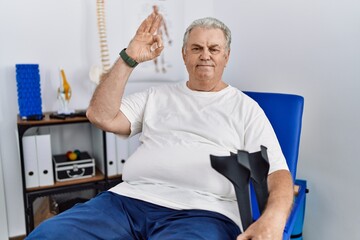 Senior caucasian man at physiotherapy clinic holding crutches smiling positive doing ok sign with hand and fingers. successful expression.