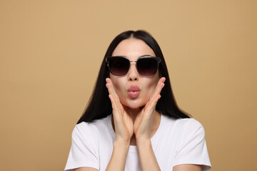 Beautiful young woman in stylish sunglasses blowing kiss on beige background