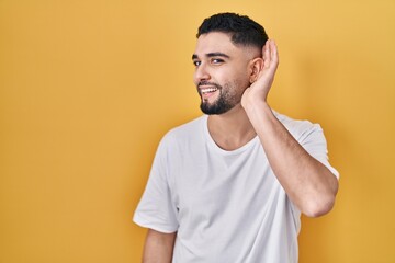 Fototapeta na wymiar Young handsome man wearing casual t shirt over yellow background smiling with hand over ear listening an hearing to rumor or gossip. deafness concept.