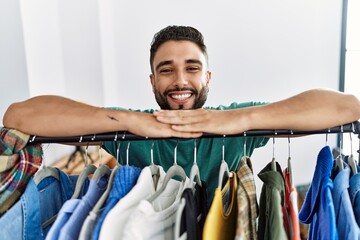 Young arab man customer smiling confident leaning on rack at clothing store