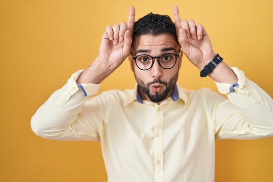 Hispanic young man wearing business clothes and glasses doing funny gesture with finger over head as bull horns