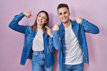 Young hispanic couple standing over pink background smiling cheerful showing and pointing with fingers teeth and mouth. dental health concept.