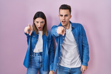 Young hispanic couple standing over pink background pointing displeased and frustrated to the...