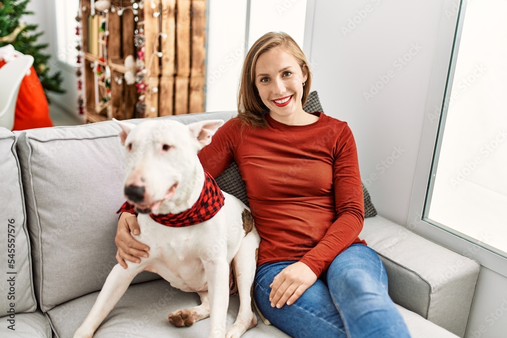 Sticker young caucasian woman smiling confident hugging dog sitting by christmas tree at home