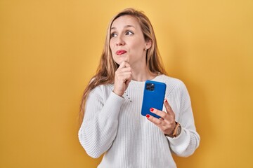 Young blonde woman using smartphone typing message with hand on chin thinking about question, pensive expression. smiling and thoughtful face. doubt concept.