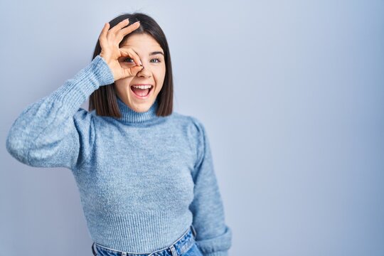 Young hispanic woman standing over blue background doing ok gesture with hand smiling, eye looking through fingers with happy face.