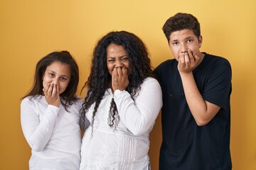 Family of mother, daughter and son standing over yellow background laughing and embarrassed giggle...