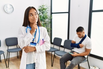 Obraz premium Young asian doctor woman at waiting room with a man with a broken arm with hand on chin thinking about question, pensive expression. smiling with thoughtful face. doubt concept.