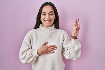 Young south asian woman standing over pink background smiling swearing with hand on chest and...