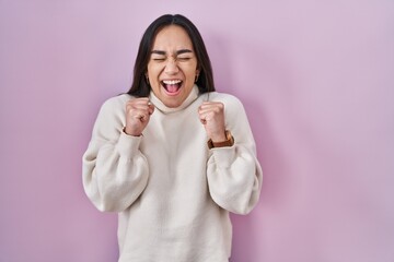 Young south asian woman standing over pink background excited for success with arms raised and eyes closed celebrating victory smiling. winner concept.