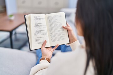 Naklejka premium Young hispanic woman reading book sitting on sofa at home
