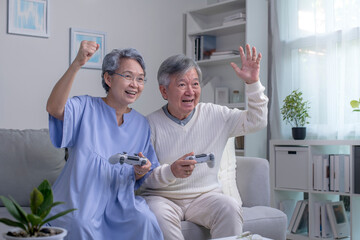 Happy Asian senior couple sitting together on their living room sofa holding controllers and laughing while playing a video game.