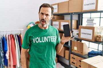 Middle age man with beard wearing volunteer t shirt holding smartphone in shock face, looking skeptical and sarcastic, surprised with open mouth