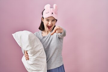 Woman with down syndrome wearing sleeping mask hugging pillow pointing displeased and frustrated to the camera, angry and furious with you