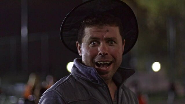 Handsome young caucasian man in a sorcerer's hat makes faces at night on the street on a blurred background of a city street with people passing by,side view close-up in slow motion.Halloween celebrat
