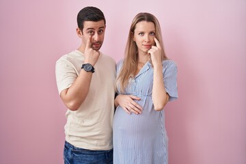 Young couple expecting a baby standing over pink background pointing to the eye watching you...
