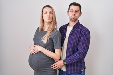 Young couple expecting a baby standing over white background puffing cheeks with funny face. mouth inflated with air, crazy expression.