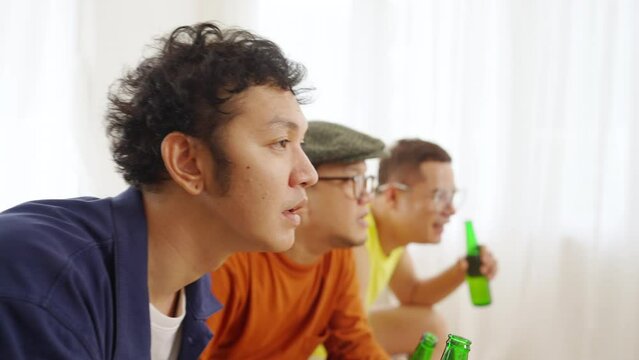 4K Group of Asian man friends watching world soccer games football competition on television with drinking beer together at home. Sport fans shouting and celebrating football team victory the match