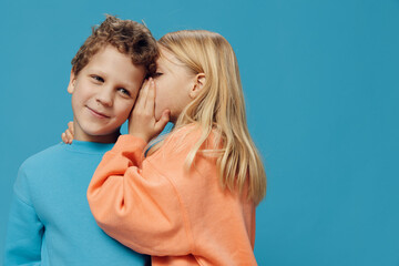  children of school age in colored sweaters stand on a blue background and the girl tells the boy a secret in the ear while standing sideways to the camera. The theme of friendship between children