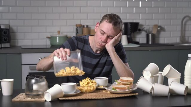 Tired man eating in the kitchen after hard day or night in messy kitchen at home. Freelance and hardworking concept