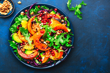 Autumn pumpkin salad with caramelazed pumpkin slices, red cabbage, avocado, arugula, pomegranate seeds and walnuts. Healthy vegan, vegetarian eating, comfort food. Blue background. Top view