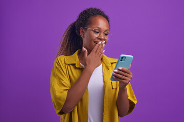 Young positive dazed African American woman closes mouth with hand looking at mobile phone pleasantly surprised and embarrassed after reading news in tabloid online press stands on lilac background - obrazy, fototapety, plakaty