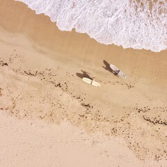 Drone shot of two surfers carrying longboards and walking on sandy beach with waves washing up on beach leaving white foam tracks. 