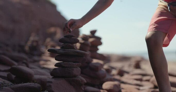 Boy finishes builidng inuksuk rock sculpture on beach with very tiny top rock
