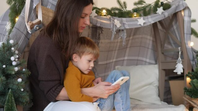 A cute little boy and his mom are reading a fairy tale on a mobile phone, comfortably ensconced in a tent in a dark night bedroom. The concept of love, childhood, friendship, imagination, creativity