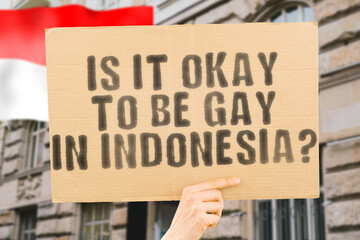The question " Is it okay to be gay in Indonesia? " is on a banner in men's hands with blurred background. Friendly. Passionate. Contact. Date. Dating. Lover. Partner. Boyfriend. Pleasant. Approval