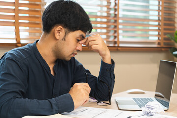 Exhausted, stressed asian young businessman, male working, took off glasses, massage nose from dry eyes, suffer on hard work while use laptop computer at office home. Overtime job, debt problem people