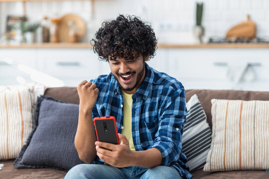 Excited young man holding mobile phone reading good news winning lottery game. Happy Indian male receives online gift feeling joy sitting at home on the couch