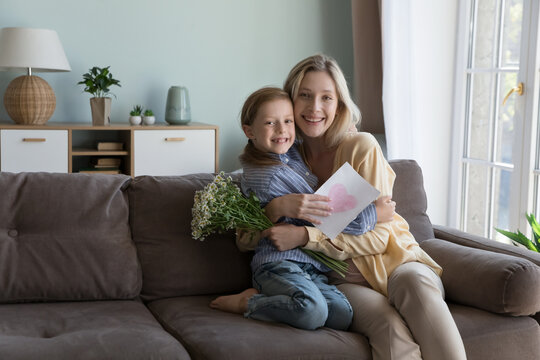 Loving family celebrating life events, Happy Mothers or International Womens Day congratulations. Little girl and young attractive mom hugging on sofa pose for picture with spring flowers and postcard