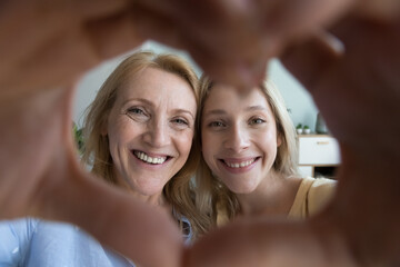 Happy mature woman and young adult daughter showing heart symbol, smile looking at camera through joined fingers make sign of love. Family ties and bond, advertise professional dental clinic services