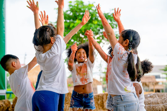 Group of different ethnic little child raise their hands or cheers together to express happiness and their clothes stain with multicolor after they play with color painting.