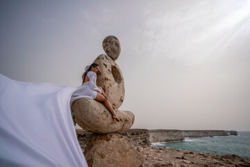 A woman sits on a stone sculpture made of large stones. She is dressed in a white long dress, against the backdrop of the sea and sky. The dress develops in the wind.