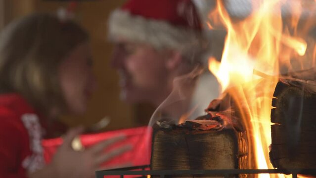 CLOSE UP: Lit fireplace and man giving a woman Christmas present in background. View of fireplace and young twosome celebrating festive winter Evening and enjoying cosy atmosphere in home living room