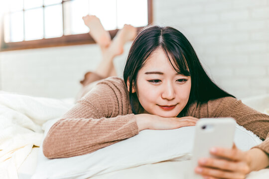 ベッドルームでパジャマを着てリラックスしながらスマホを見る笑顔の日本人女性

