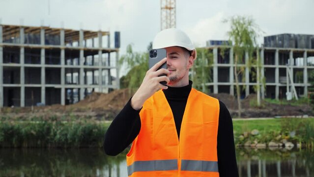 Young attractive engineer man in a white safety helmet makes photos of buildings under construction on a mobile phone. Male person sends messages to the customer via a smartphone. Engineering project.