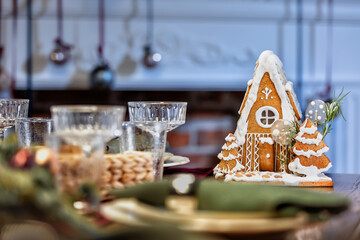 A small gingerbread house with icing, decorated for Christmas. Standing among the festive table set with festive dishes