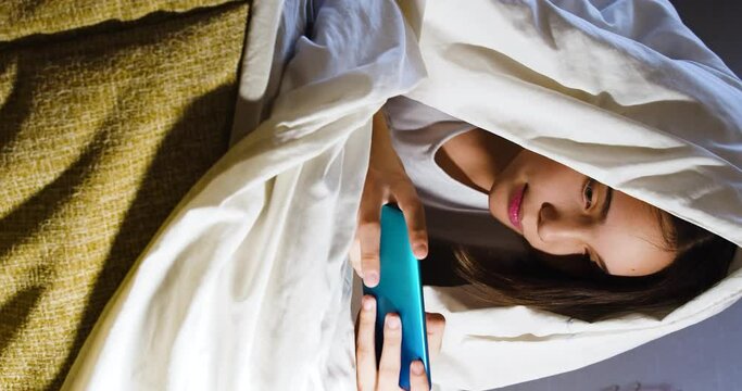 Teenager girl lies in bed with smartphone unger the blanket. Vertical video.