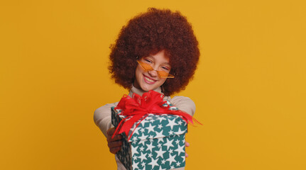 Lovely smiling young teenager child girl kid presenting birthday gift box stretches out hands, offer wrapped present career bonus, celebrating party. Little funny children on studio yellow background