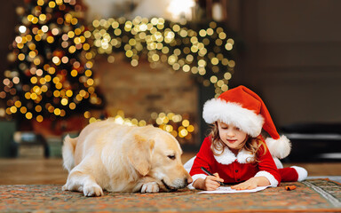 A child write letter for Santa near a her favorite dog. Festival decorated Christmas tree and fire...