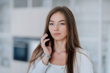 Pensive young Caucasian woman in white suite making call by phone looks aside with calm face. Successful Swedish female lawyer consulting client remote working home. Businesswoman at office.