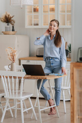 Adorable blonde young caucasian woman with ponytail in blue jeans and shirt standing at table with laptop waits for remote lesson. Swedish girl at remote study at kitchen, home. Adorable student girl.