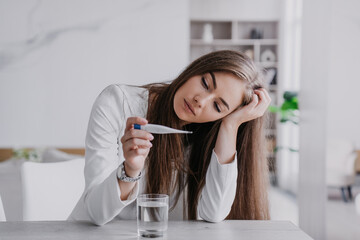 Attractive blonde caucasian young woman in white suit feels bad sitting at table holds thermometer measures body temperature. Beautiful Caucasian female leans on hand feels fatigue. Illness