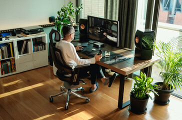 Back view of middle aged man working on his computer at home by a window at modern workplace in room decorated with green potted plants. Graphic designer working from home