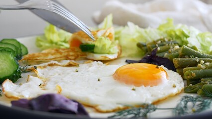 Fried eggs with stewed vegetables and greens are tasty and nutritious. Breakfast on a keto diet is rich in fats and proteins. On a fried egg, greens and cucumber are folded with a fork and knife.